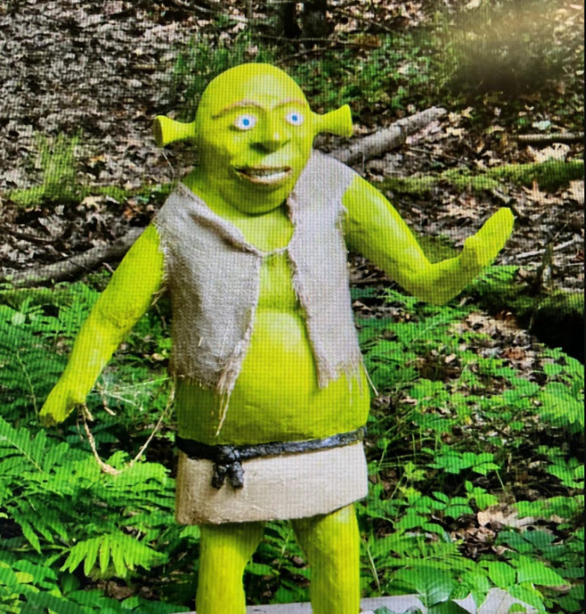 Low-res image of handmade Shrek statue showing a bright green ogre with funnel-like ears and blue eyes wearing a burlap vest with one hand extended as in a wave.