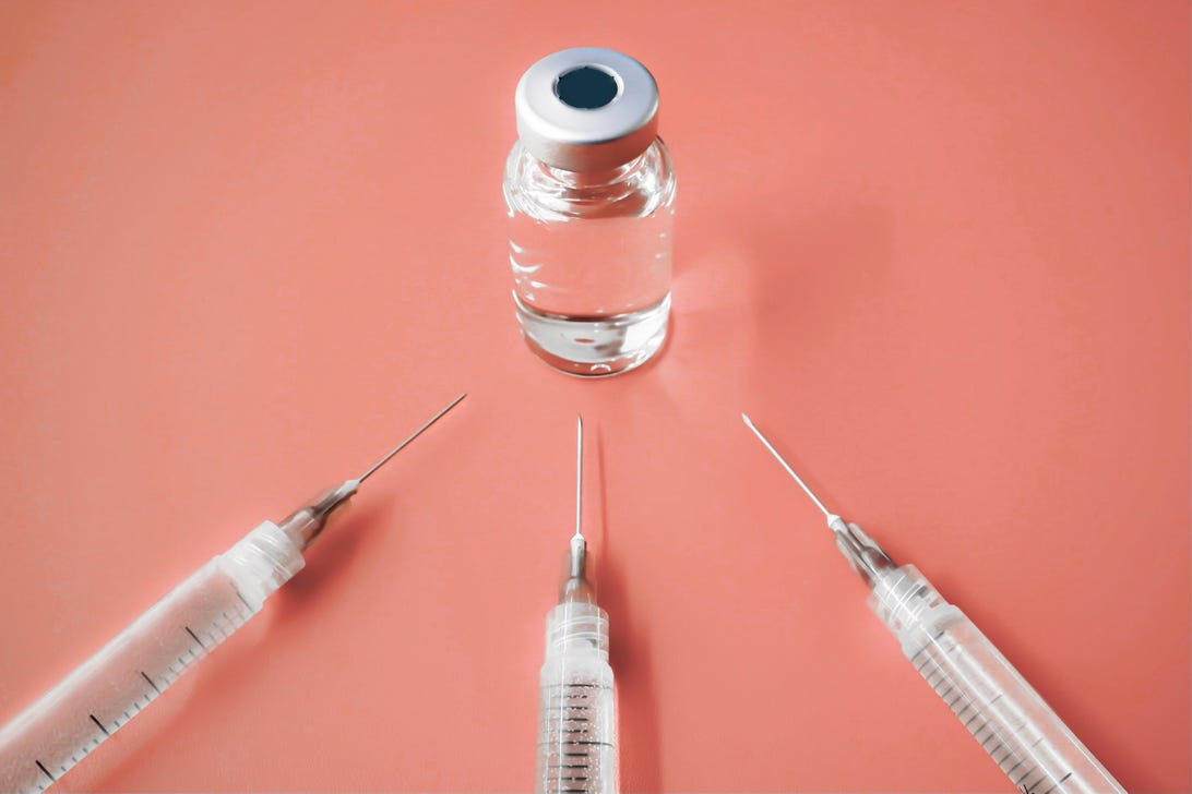 A COVID-19 vaccine vial and three needles against a pink background.