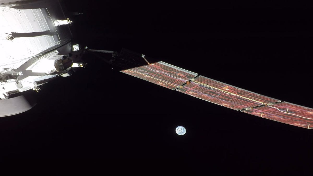Part of the white Orion spacecraft and an extended solar array against black space with the partially lit Earth looking dainty.
