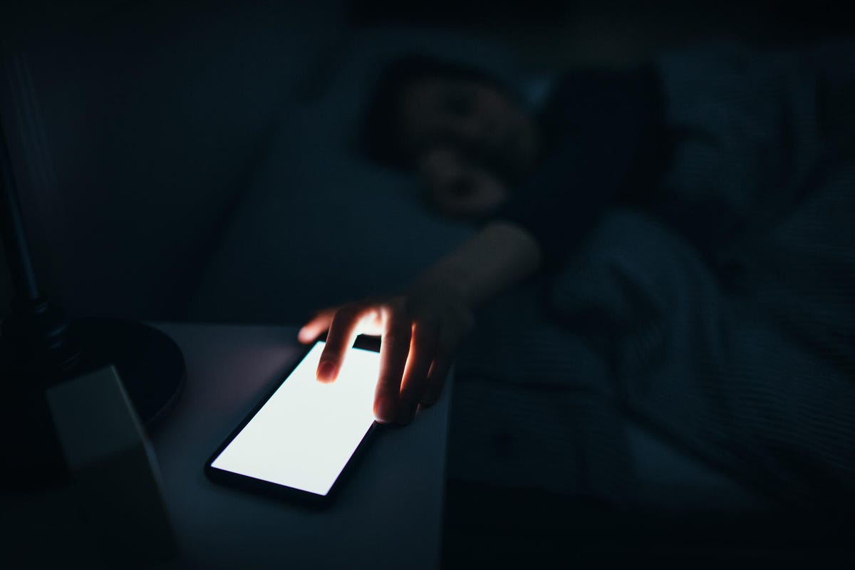 Dans une pièce sombre, une personne sort d'un lit pour un téléphone avec un écran très éclairé