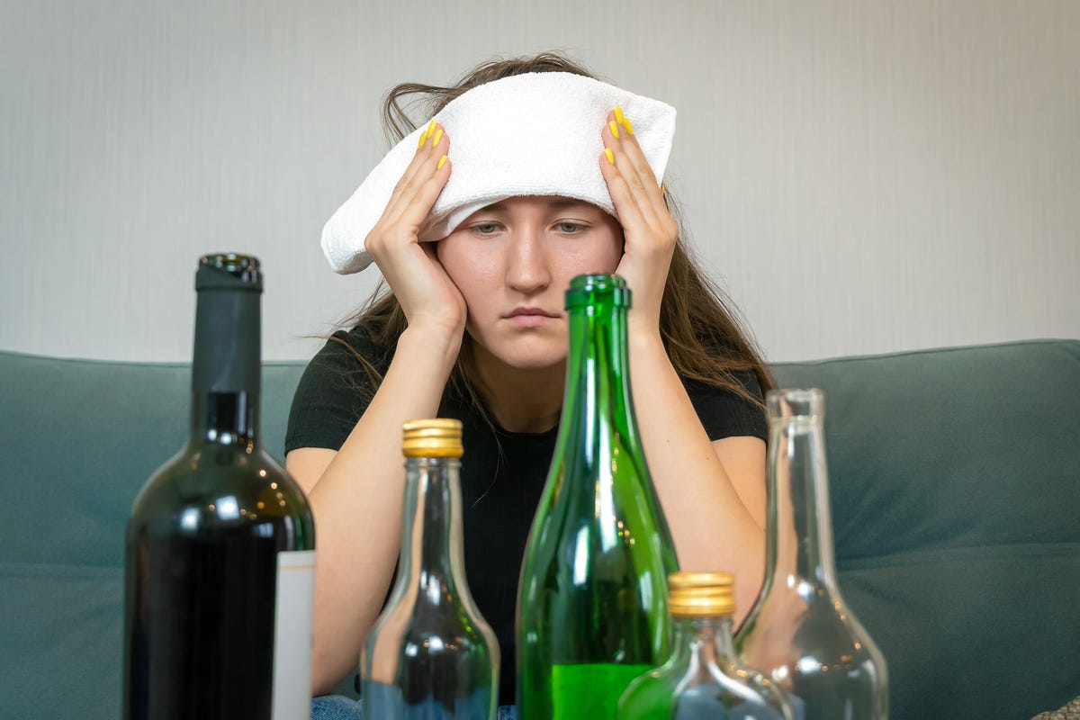 Young woman with towel on her forehead, starting at empty bottles of alcohol.