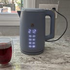 The Beautiful by Drew Barrymore One-Touch Electric Kettle sits on a granite countertop beside an inviting cup of tea.
