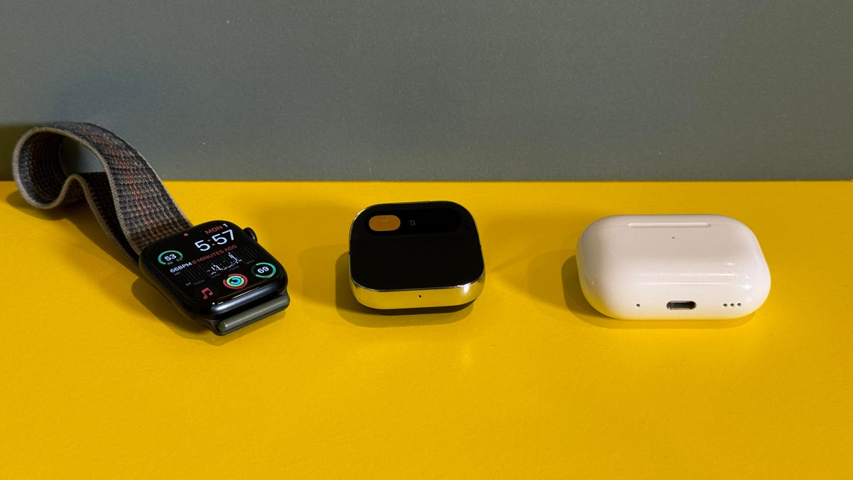 Humane AI Pin on a yellow table between an Apple Watch and AirPods case
