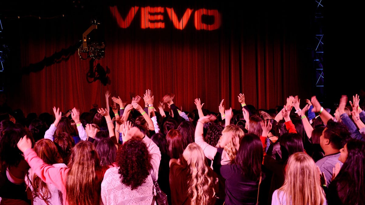 A crowd applauds in front of a stage with a velvet curtain where VEVO is projected in white light.