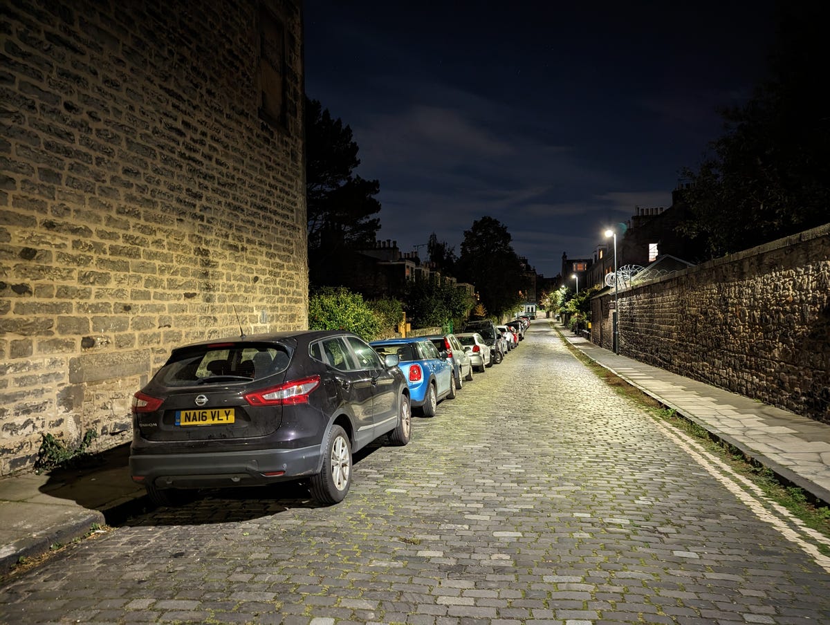 An image showing a night-time street with cars parked on the left.