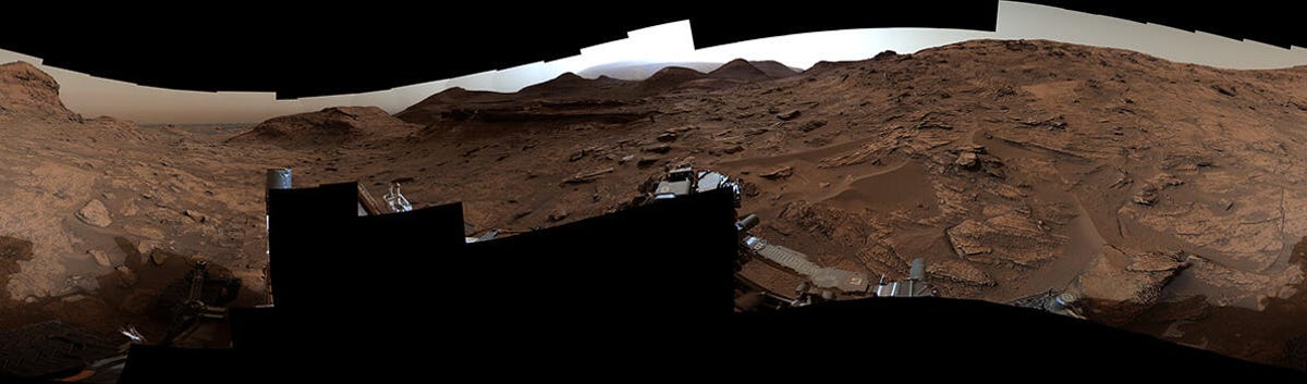 A dark, craggy Mars landscape show part of the Curiosity rover at the bottom and a stretch of reddish-brown hills in the distance with rock and boulders closer to the camera.