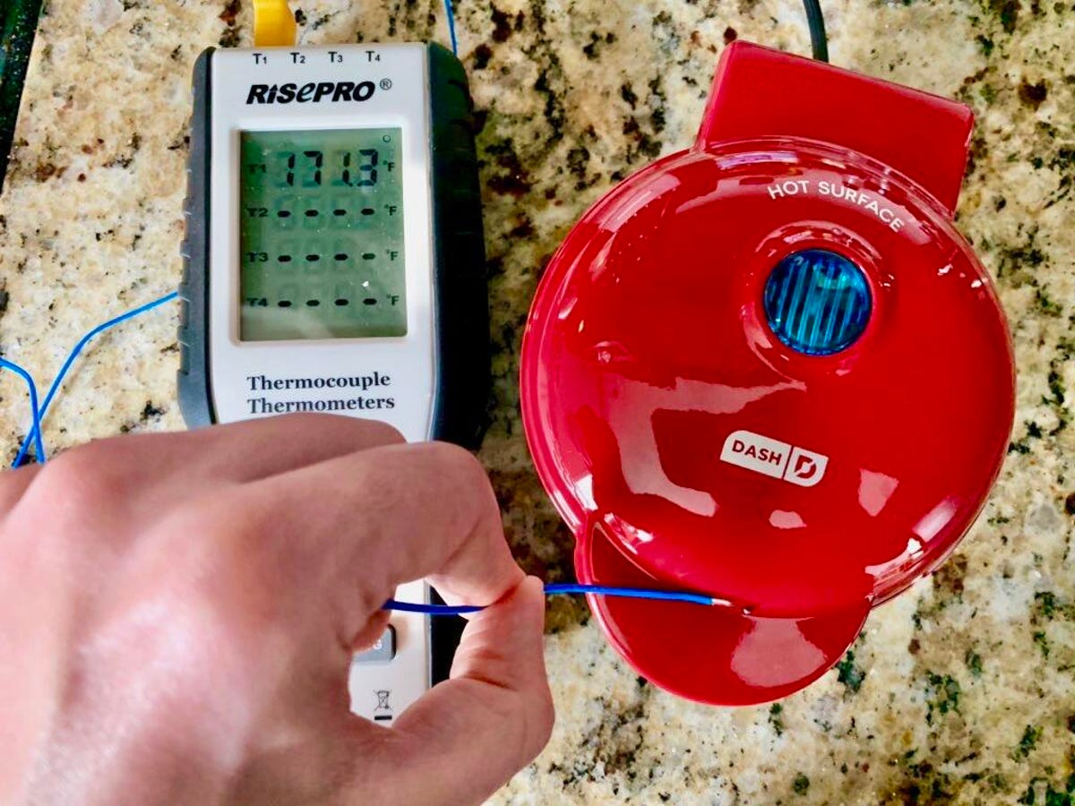 A hand using a digital thermostat to measure the temperature of a red Dash waffle maker.