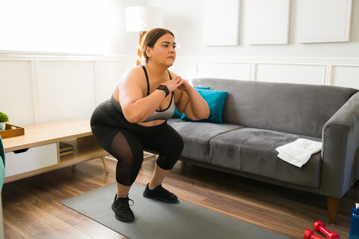 Young midsize woman does squats on a yoga mat in living room.