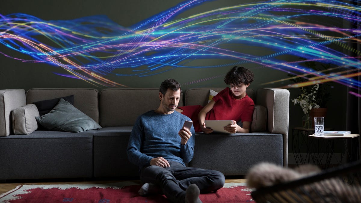 A man and woman sit or lean on a couch using their devices while colorful threads representing Wi-Fi internet float above them.