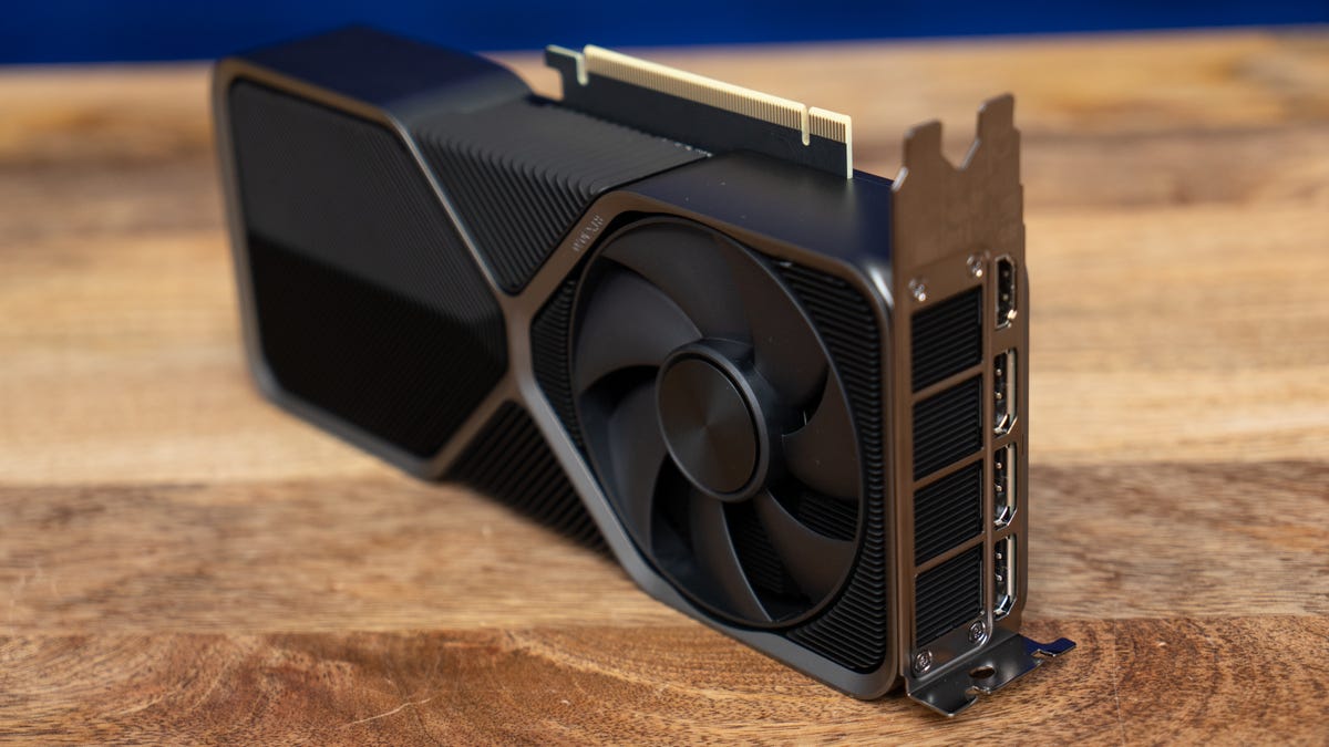 The RTX 4070 FE facing forward, back side facing you, angled up to your right showing the connectors and rear vent, on a wood surface