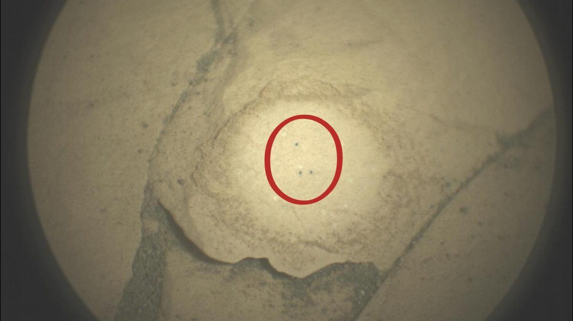 A close-up look at three tiny L-shaped dots on a beige Martian rock surrounded by a red circle.