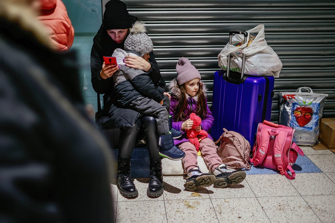 Ukrainian refugees at an aid center in Poland. A woman all in black holds a girl on her lap while peering intently at a red smartphone.