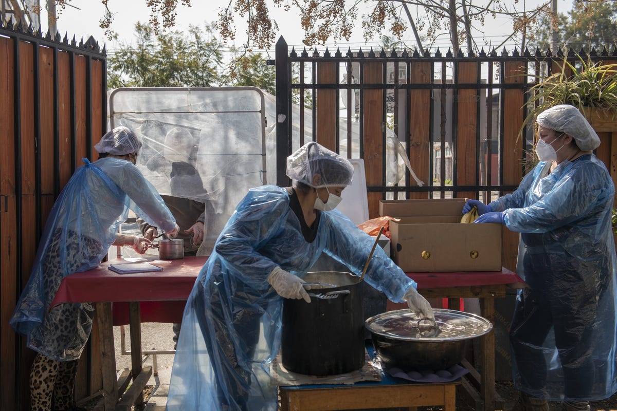 Three people in hairnets and pale blue uniforms prepare food behind a fence to deliver to those in need.
