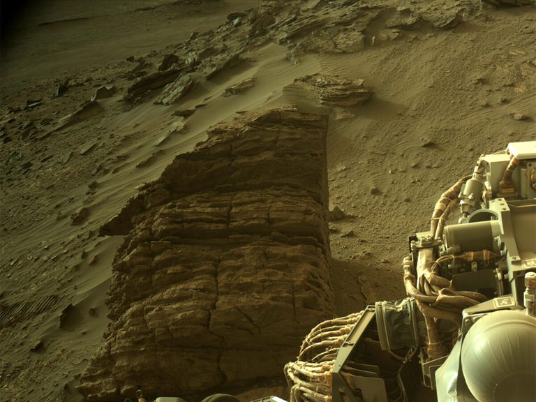 A layered rock sits on the Mars surface with part of the rover visible in the corner.