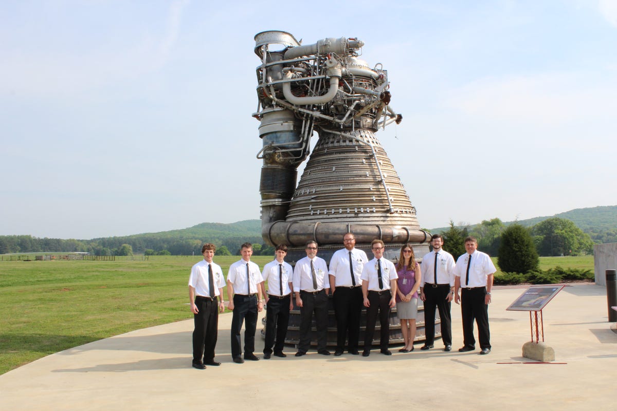 A group of engineers dressed in Apollo-era style stand in front of a massive rocket engine outside.