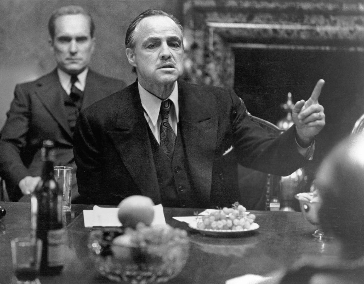 American actor Marlon Brando (1924 - 2004), in character as mob kingpin Don Vito Corleone, gestures as he sits at a table as colleague and compatriot Robert Duvall, as Tommy Hagen, sits behind him in a scene from the gangster film 'The Godfather,' directed by Francis Ford Coppola, 1972. (Photo by Paramount Studios courtesy of Getty Images)
