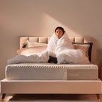 The Casper Nova Hybrid mattress on a light-colored bed frame, with a woman wrapped in a blanket on top.