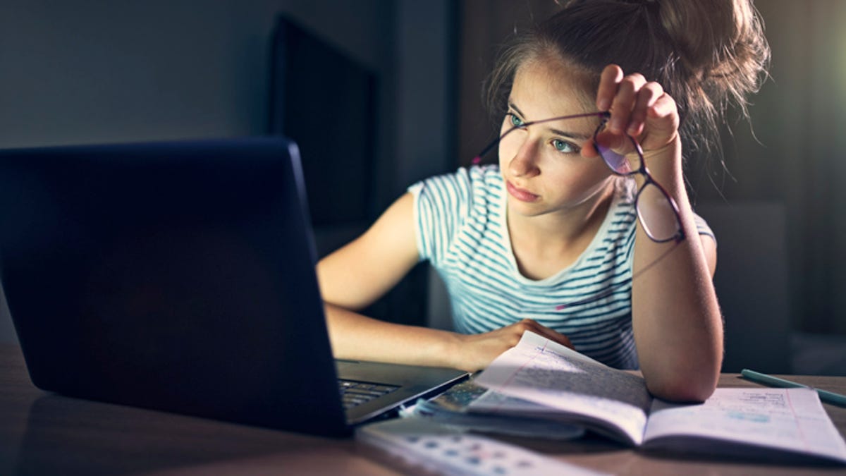 Tired woman sitting in front of her laptop, with her arm propped on her books, holding her glasses