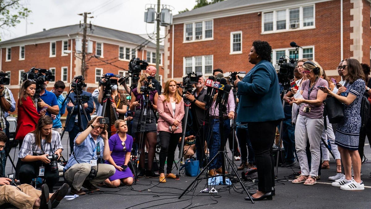 Georgia gubernatorial candidate Stacey Abrams speaks in front of a circle of reporters, some standing with big cameras and others crouching while recording with smartphones..