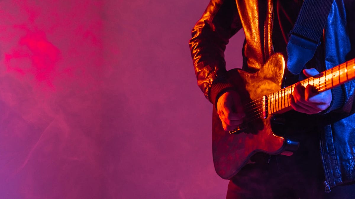 A person in a leather jacket plays the guitar against a fuchsia background