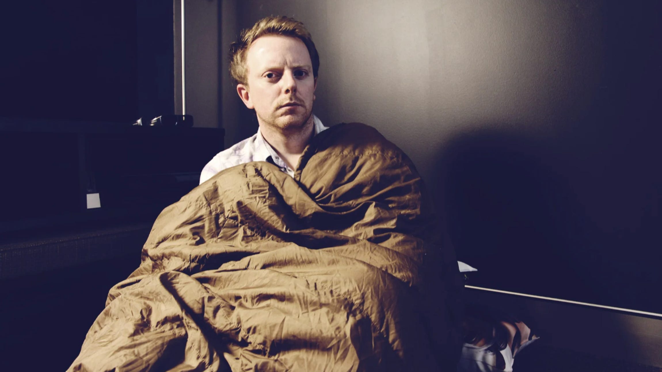 A somewhat dazed- and paranoid-looking Mark Serrels peers at the camera while sitting up out of a sleeping bag, which he clutches to his chest.