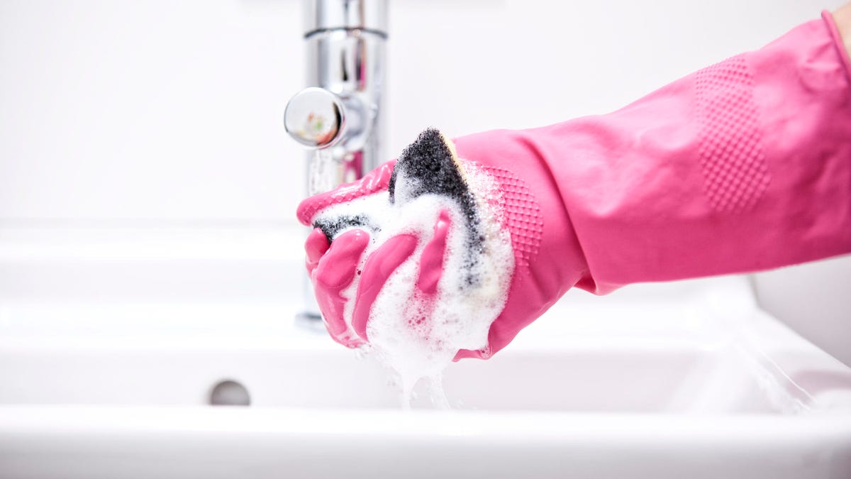 Above a sink, a hand in a pink rubber glove squeezes soap from a sponge