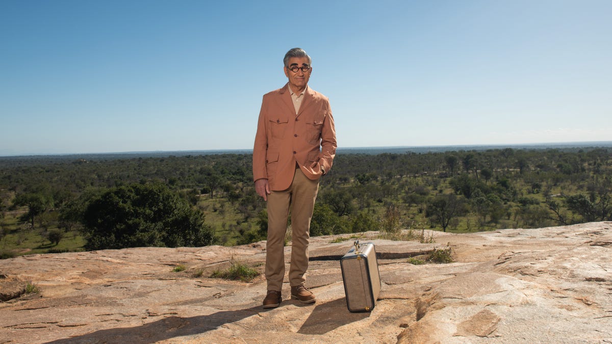 Eugene Levy stands next to a suitcase.
