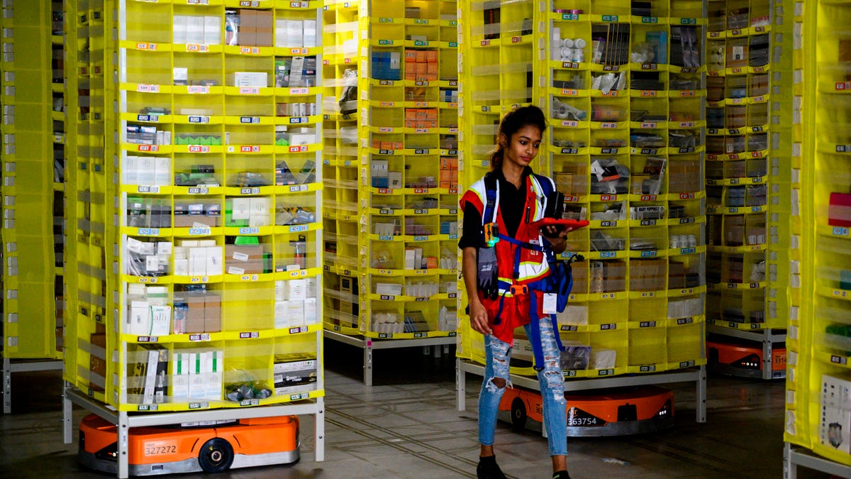 A worker at an Amazon fulfillment center in Staten Island holds a tablet for controlling robots. Behind her are bright yellow shelves filled with packaged goods.