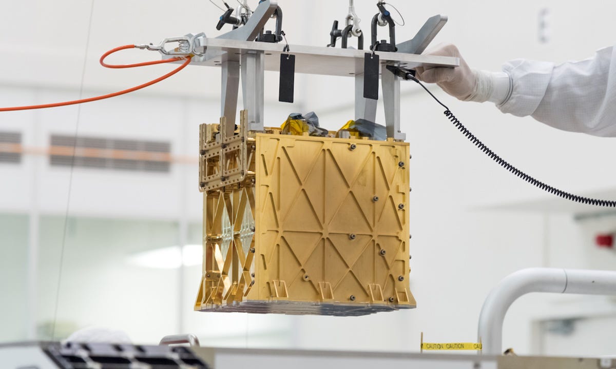 In what looks like a laboratory clean room, we can see the golden, patterned Moxie mechanism attached to wires and other metal apparatuses that are holding it up.