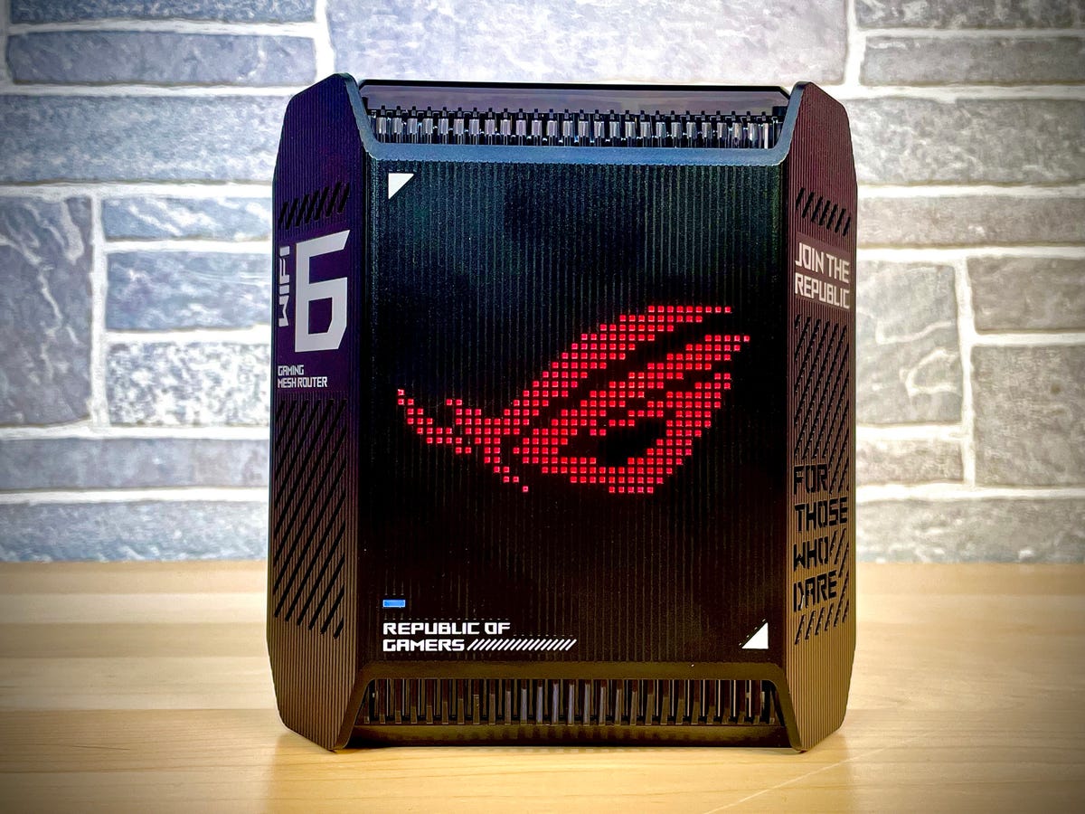 A single Asus Rog Rapture GT6 mesh gaming router sits on a wooden table. It's a black, angular device with internal antennas and a color-changing LED dot matrix logo on the front.