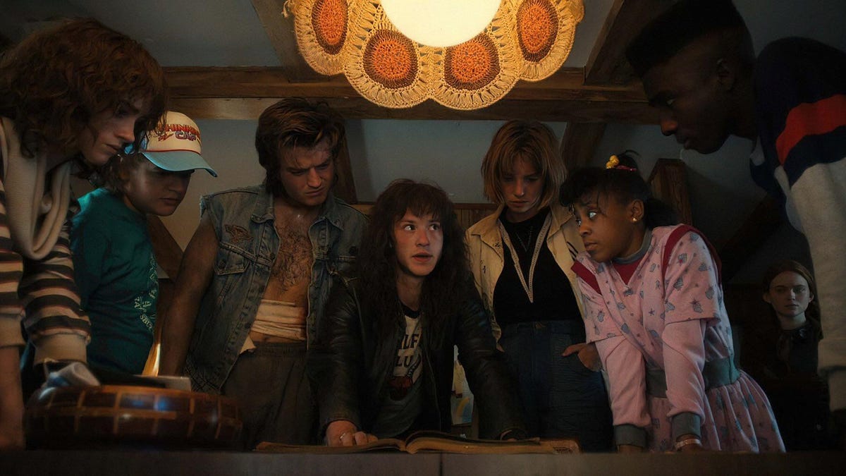 Stranger Things characters gather around a table: Nancy, Dustin, Steve, Eddie, Robin, Erica and Lucas, with Max in the background