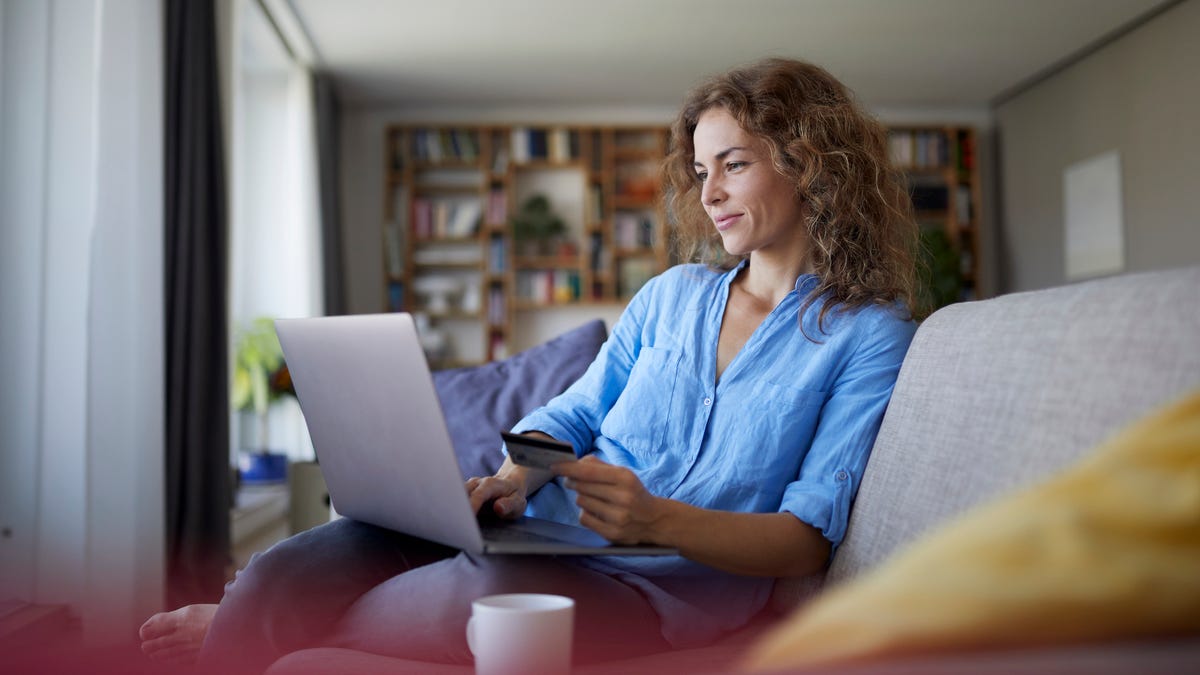 A woman wearing a blue blouse is sitting on a couch looking at her laptop with a bank card in hand.