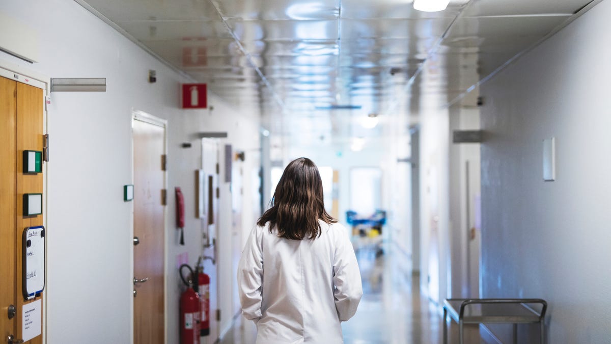 A woman in white scrubs walks through a hospital hallway.