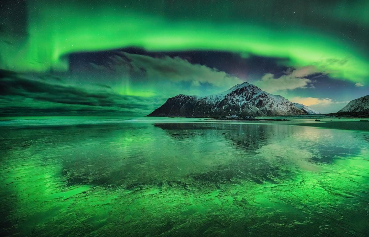 Circle of green aurora light from the sky reflects in water below with a snowy mountain peak in the middle.