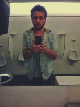 A disheveled man takes a selfie in a men's room, with urinals in the background