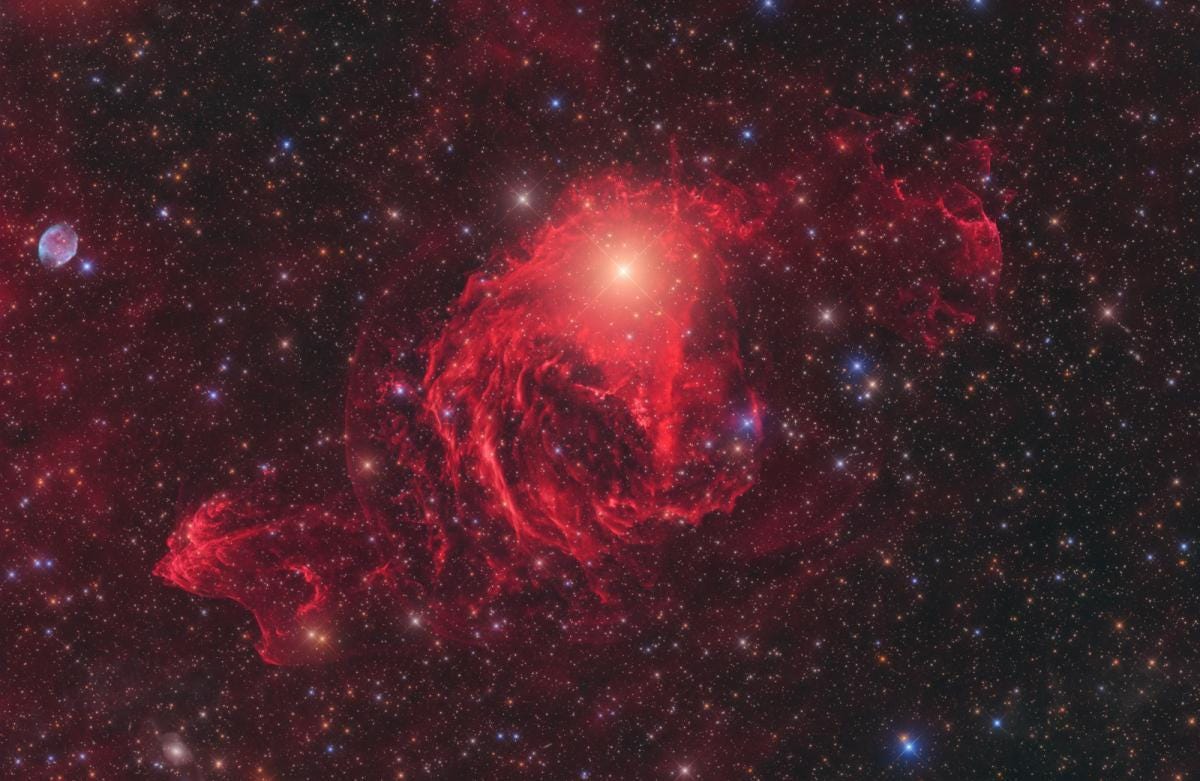 Red, cloud-like nebula glows against a reddish and black backdrop of stars.