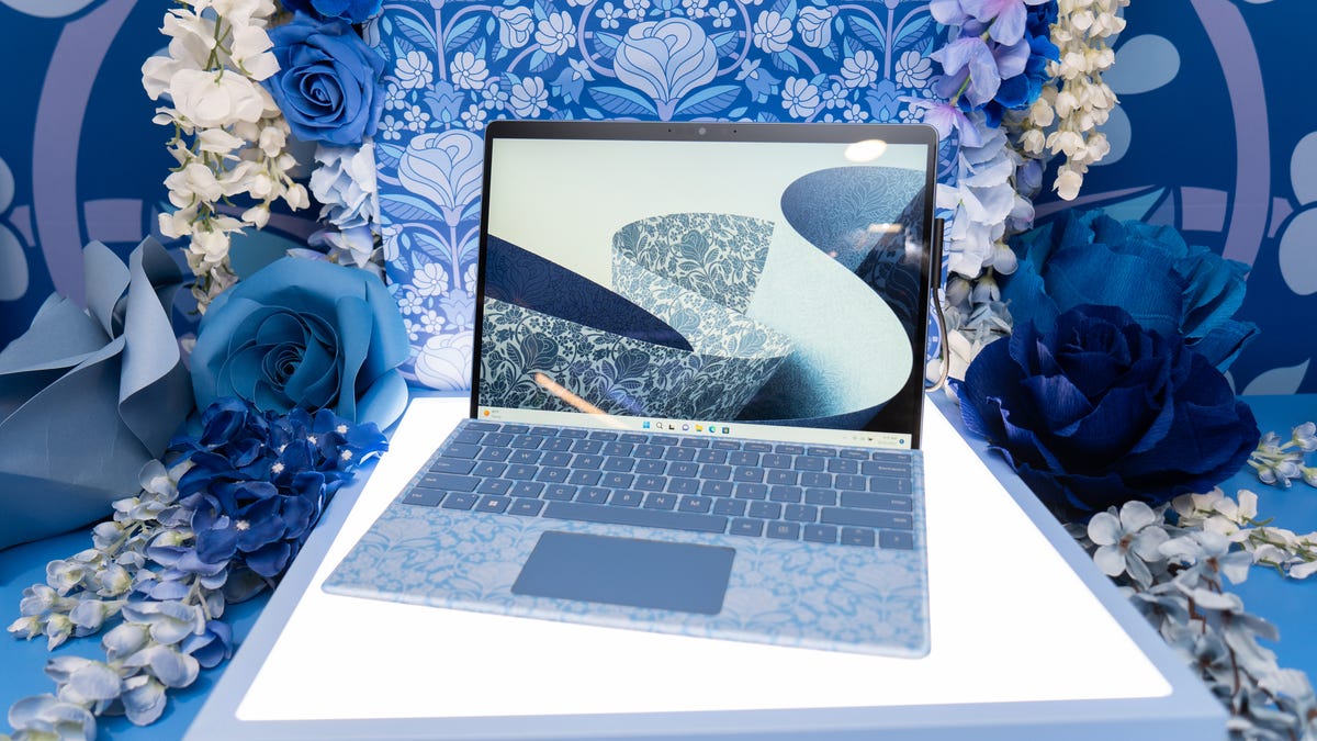 Microsoft Surface Pro 9 Liberty Edition poised with matching keyboard on a light table surrounded by a complementary backdrop and fake flowers.