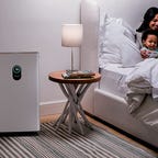 A woman reads a book to a little boy as they lie in bed and the Shark HE601 Air Purifier 6 rests nearby.